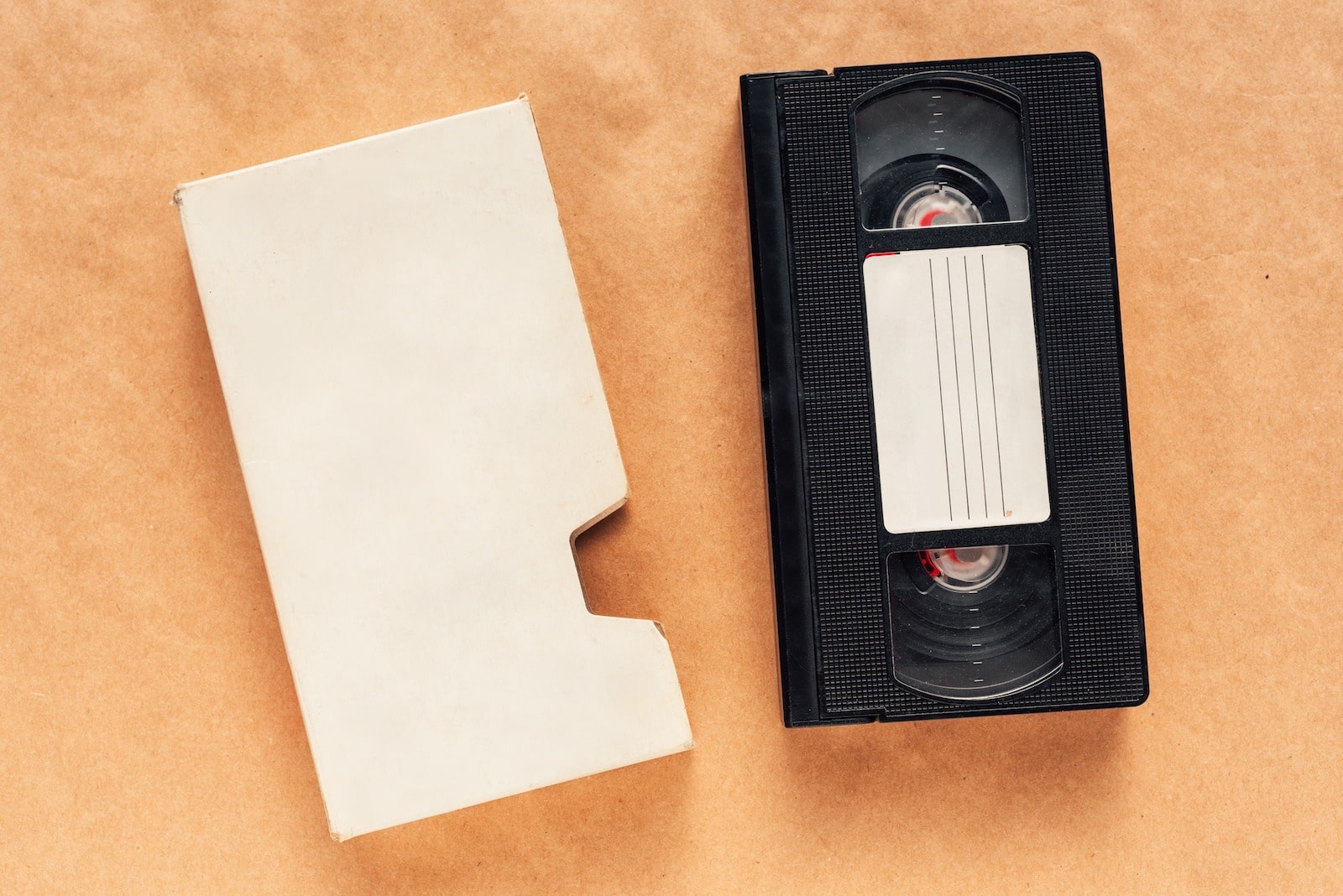 A VHS tape with a blank label next to an empty white cardboard sleeve on a brown surface.