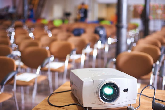 A projector on a table in a conference room with rows of empty brown chairs in the background.
